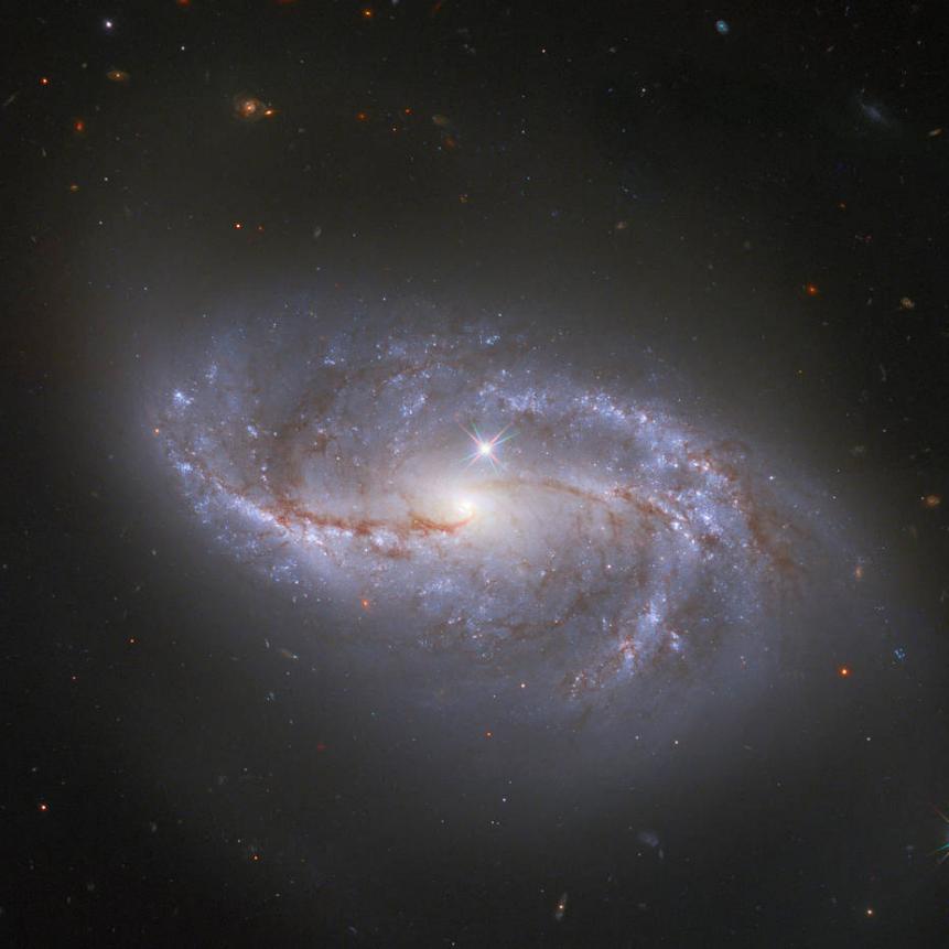 Looking deep into the Universe, the NASA/ESA Hubble Space Telescope catches a passing glimpse of the numerous arm-like structures that sweep around this barred spiral galaxy, known as NGC 2608. Appearing as a slightly stretched, smaller version of our Milky Way, the peppered blue and red spiral arms are anchored together by the prominent horizontal central bar of the galaxy. In Hubble photos, bright Milky Way stars will sometimes appear as pinpoints of light with prominent lens flares. A star with these features is seen in the lower right corner of the image, and another can be spotted just above the pale centre of the galaxy. The majority of the fainter points around NGC 2608, however, lack these features, and upon closer inspection they are revealed to be thousands of distant galaxies. NGC 2608 is just one among an uncountable number of kindred structures. Similar expanses of galaxies can be observed in other Hubble images such as the Hubble Deep Field which recorded over 3000 galaxies in one field of view.