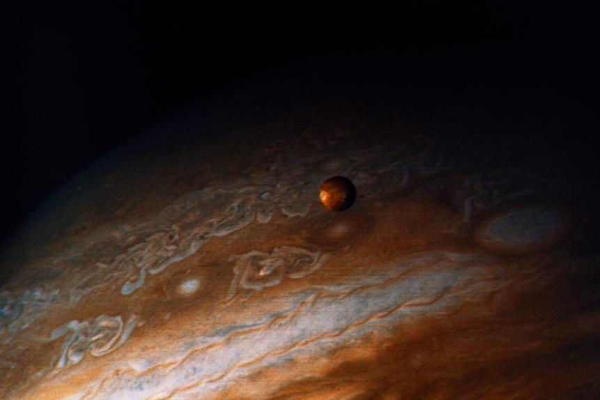 (Original Caption) This photograph of the southern hemisphere of Jupiter was obtained by Voyager 2 on June 25, 1979, at a distance of 12 million kilometers, (8 million miles). The Voyager spacecraft is rapidly nearing the giant planet, with closest approach to occur at 4:23 pm PDT on July 9th. Seen in front of the turbulent clouds of the planet is Io, the innermost of the large Galilean satellites of Jupiter. Io is the size of our moon. Voyager discovered in early March that Io is the most volcanically active planetary body known in the solar system, with continuous eruptions much larger than any that take place on the Earth. The red, orange, and yellow colors of Io are thought to be deposits of sulfur and sulfur compounds produced in these eruptions. The smallest features in either Jupiter or Io can be distinguished in this picture are about 200 kilometers (125 miles) across