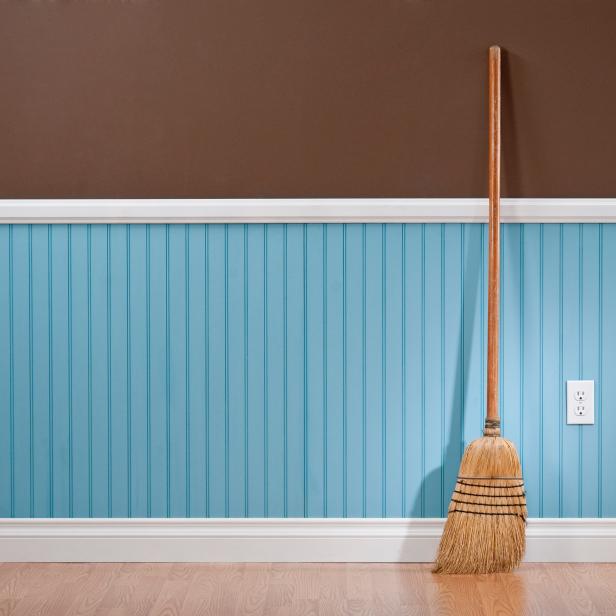 A wooden broom propped up against a wall.  The wall is two different colors.  The top half of the wall is solid chocolate brown.  The bottom half of the wall is bead board, and it has repeating parallel lines spaced a few inches apart.  The bottom section of the wall is a light shade of blue.  Propped up against the wall is a tan wooden broom.  The broom has angled bristles gathered with a dark thread.  There is white molding separating the two sections of the wall and at the floor, which is comprised of light-colored wood.