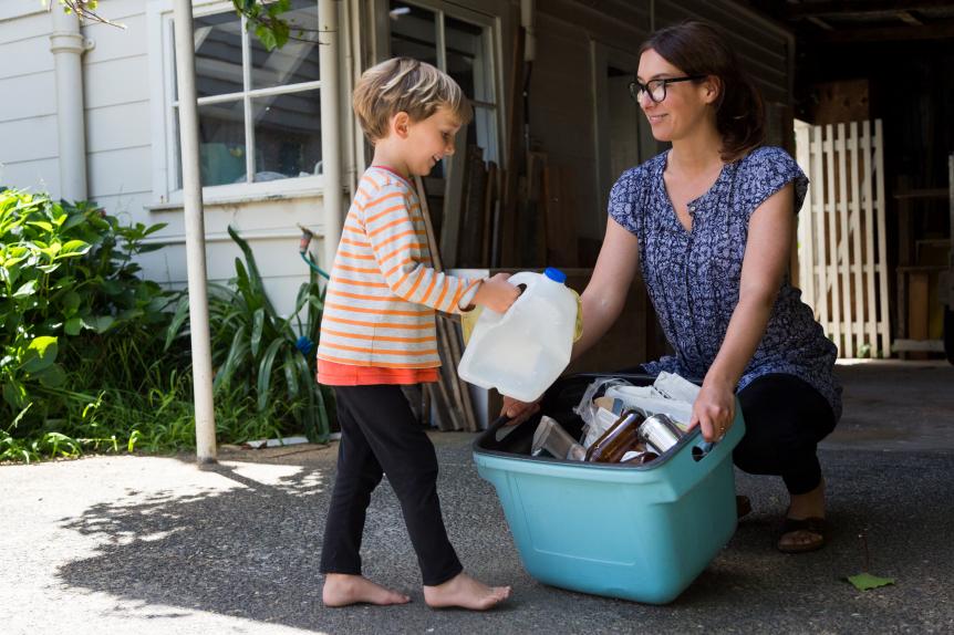 A mother and child are outside sorting through the recycling bins with the garage in the background