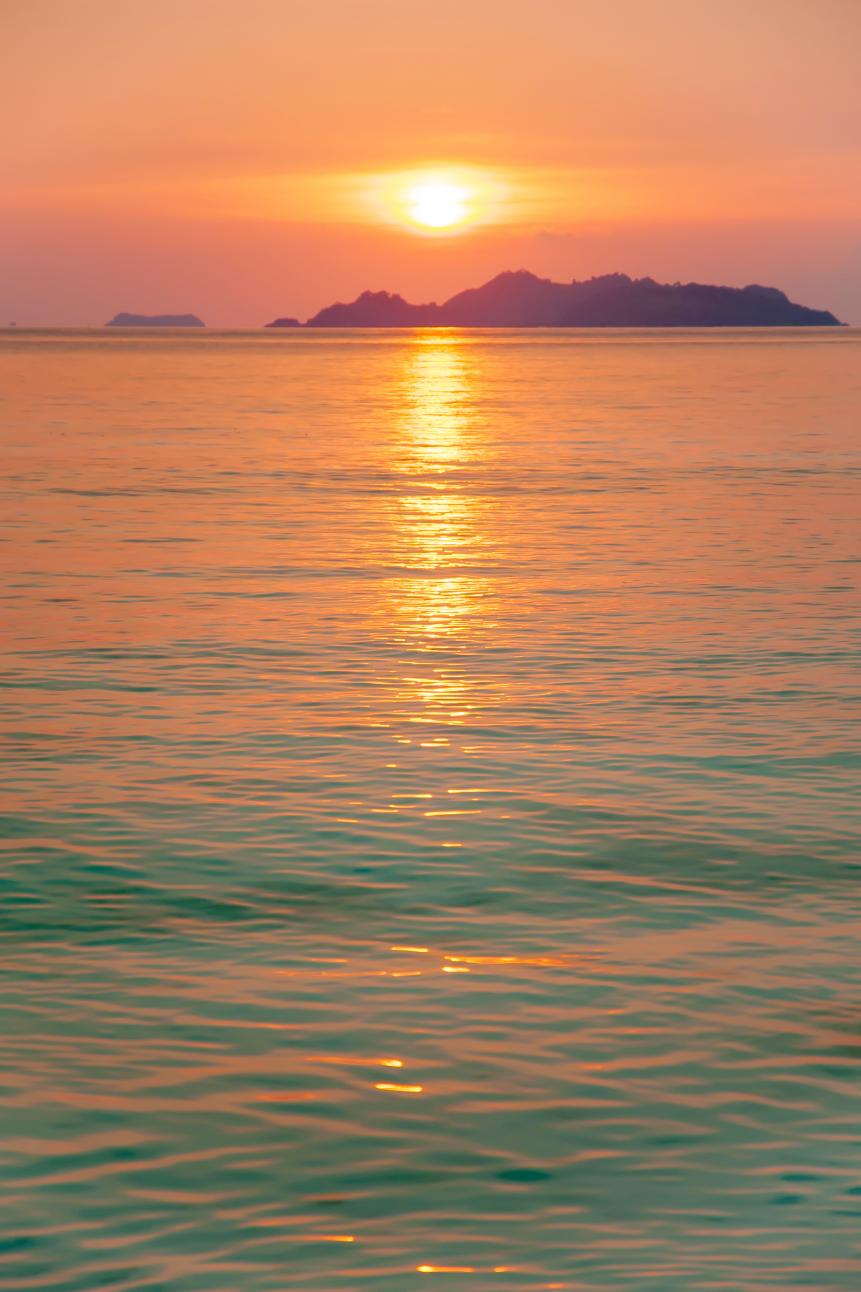 A tranquil seascape at sunset, glowing sun reflection on gently waves, unknown remote island blurred in the background. Tanintharyi, South Myanmar. Selective focus.