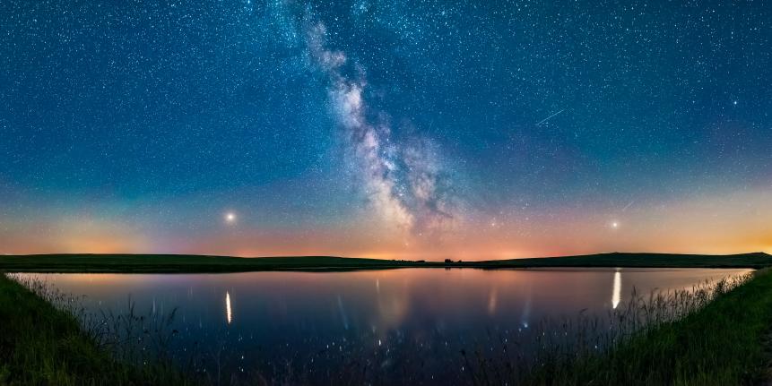 A 160 degree panorama taken July 5/6 of the summer Milky Way and the array of summer 2018 planets over a prairie pond in southern Alberta, Canada.  Mars is bright to the left, Saturn is dimmer and at centre in the Milky Way, while bright Jupiter is at right. Mars and Jupiter nicely flank the Milky Way, and cast glitter paths on the water. The arcing line joining the planets defines the arc of the ecliptic, always low in the south in northern hemisphere summer. Mars was approaching Earth and brightening at this time heading for a late July opposition.  The sky is deep blue with solstice twilight. Several satellite trails punctuate the sky.