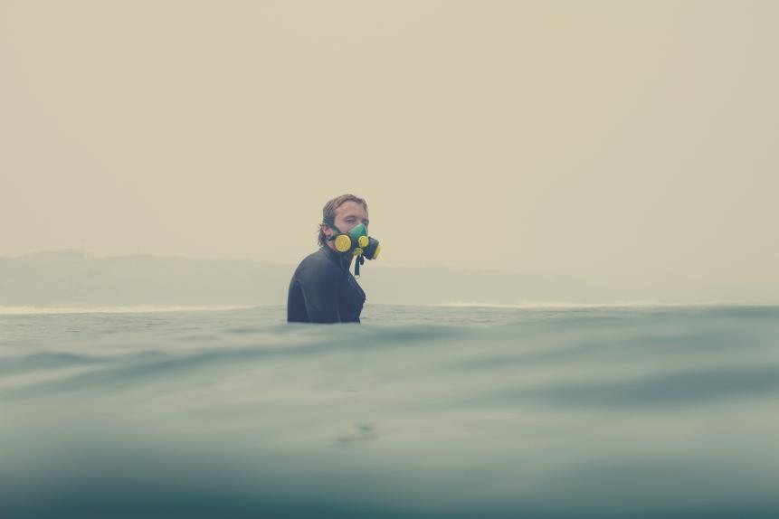 A surfer sitting out at sea and wearing a respirator due to the thick smoke haze caused by the Australian bushfires 2020. He is looking towards camera and the coastline can be made out through the thick smoke.