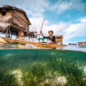 Split wide angle shot of a Bajau woman rowing wooden canoe in floating village carrying her child, showing the ocean floor and the wooden floating houses at the background, with blue sky and white clouds