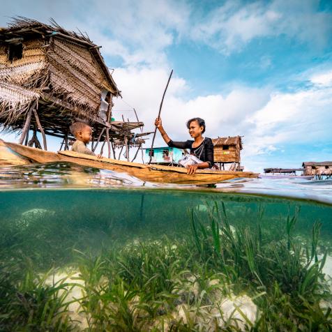 Split wide angle shot of a Bajau woman rowing wooden canoe in floating village carrying her child, showing the ocean floor and the wooden floating houses at the background, with blue sky and white clouds