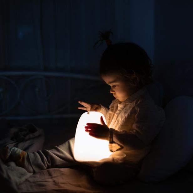 Adorable baby girl playing with bedside lamp in nursery. Happy kid sitting on bed with nightlight. Little child at home in the evening before sleep.
