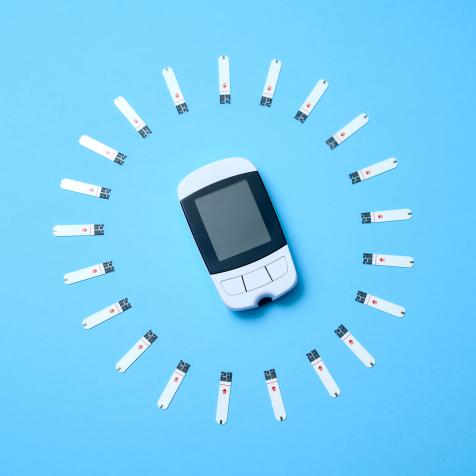 Flat lay glucometer with test stripes organised into circular pattern around device on blue background.