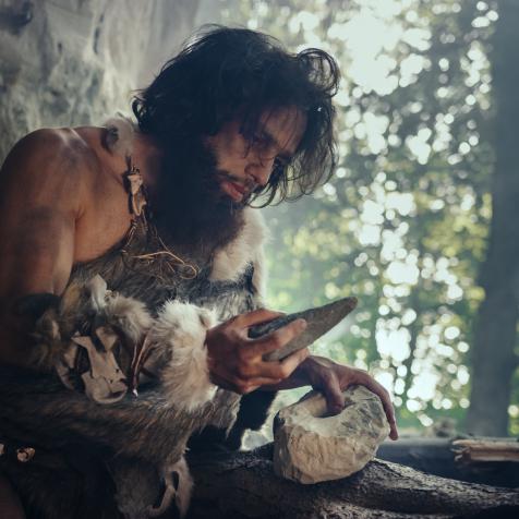Primeval Caveman Wearing Animal Skin Holds Sharp Stone and Makes First Primitive Tool for Hunting Animal Prey, or to Handle Hides. Neanderthal Using Handax. Dawn of Human Civilization
